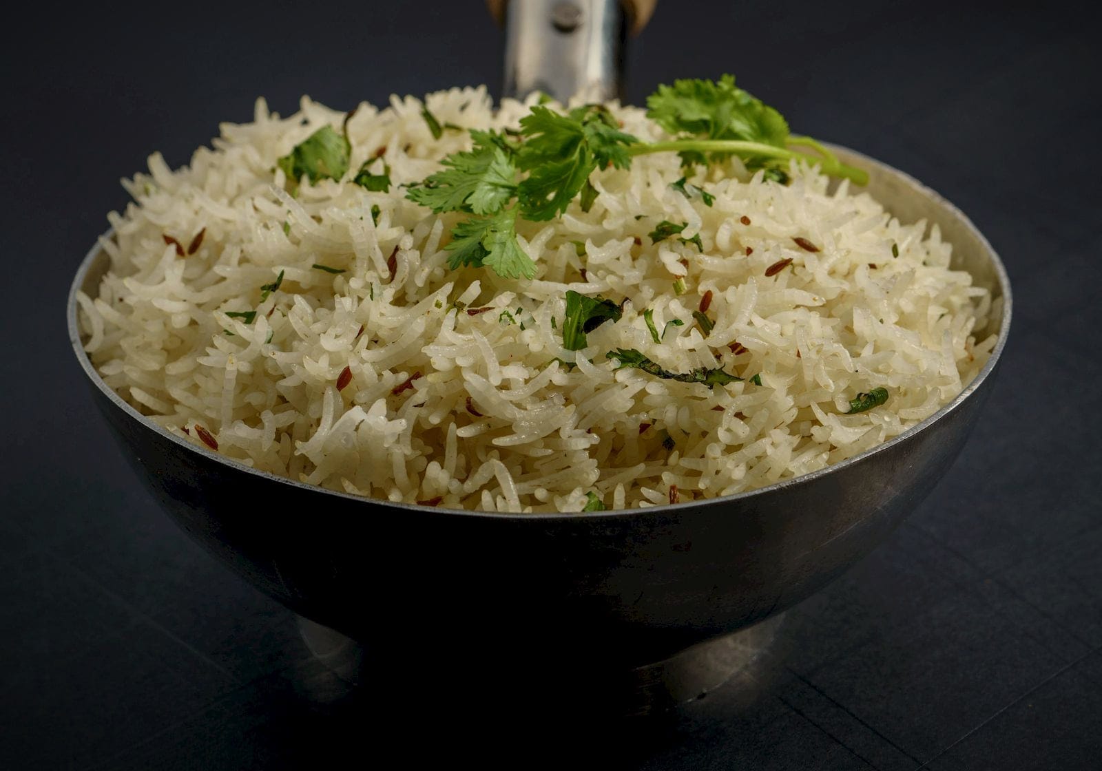 Cumin-flecked basmati rice with coriander in a steel bowl on dark slate