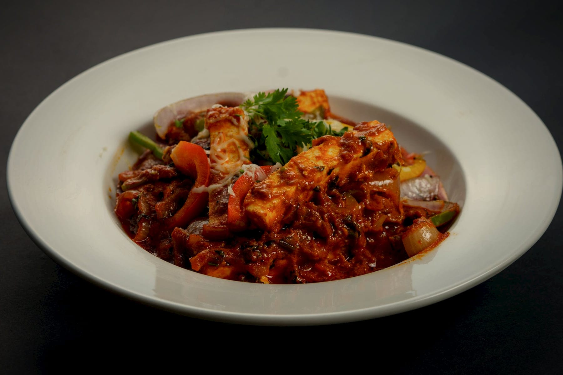 Paneer with onions and peppers in a dark, textured masala, in a wide ivory bowl