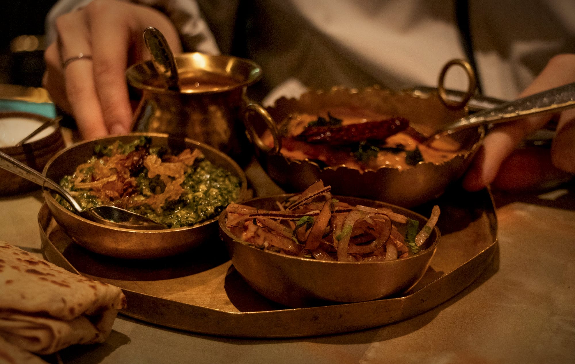 Hands serving from brass bowls of curry and dal on a brass tray, with bread at the edge of the table