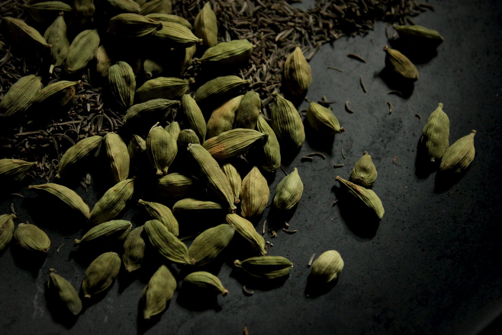 Green cardamom pods and cumin scattered on dark stone